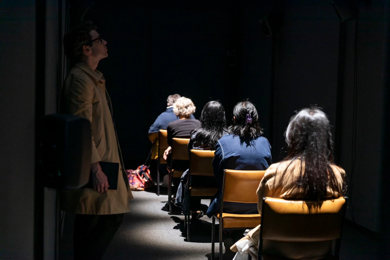 Five people sit single file in a line of padded yellow chairs, facing away into the darkness of a narrow room. The back of their heads glow under overhead lights. One person stands to the side, outside of the row of light. They look up and into the shadows of the ceiling, as though listening attentively.